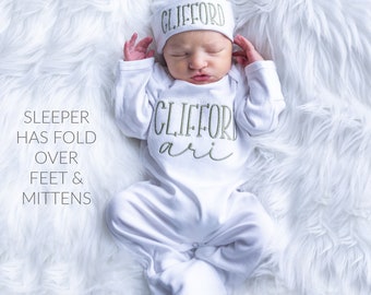 May include: White baby sleeper with fold-over feet and mittens. The sleeper has green embroidery that says "Clifford Ari". The baby is wearing a white hat with green embroidery that says "Clifford".