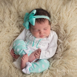 May include: A newborn baby girl wearing a white onesie with the name "Makenna" embroidered on it. She is also wearing mint green and white polka dot leggings with a white bow on each leg. The baby is sleeping on a white fluffy rug.
