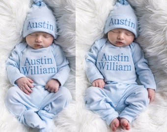 May include: A light blue baby onesie with the name "Austin William" embroidered on the front. The onesie has fold-over hands and feet. The baby is wearing a matching light blue hat with the name "Austin" embroidered on it.