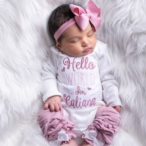 May include: A baby girl wearing a white onesie with pink embroidery that says "Hello World I'm Caliana". The baby is wearing a pink headband with a large pink bow and pink ruffled leg warmers with white bows.