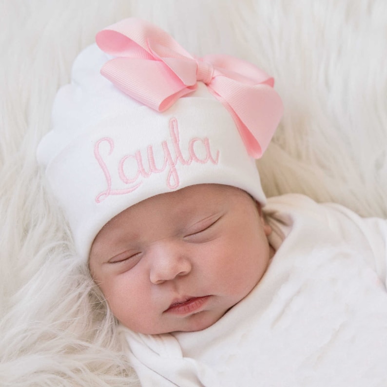 Op de afbeelding: Een witte baby muts met een roze strik en de naam "Layla" geborduurd op de voorkant. De muts wordt gedragen door een slapende baby.