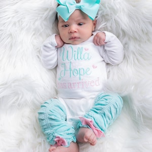 May include: A baby girl wearing a white onesie with the text "Willa Hope has arrived" in turquoise and pink. She is wearing a turquoise bow headband and matching turquoise leg warmers with pink bows.