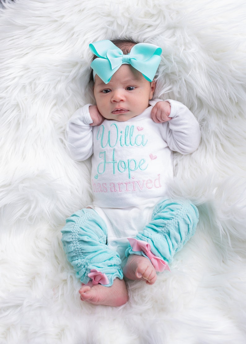 May include: A baby girl wearing a white onesie with the text "Willa Hope has arrived" in turquoise and pink. She is wearing a turquoise bow headband and matching turquoise leg warmers with pink bows.