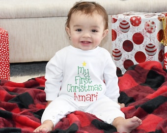 May include: A baby wearing a white onesie with the text "My First Christmas Amari" in green and red, with a Christmas tree graphic. The baby is sitting on a red and black plaid blanket.
