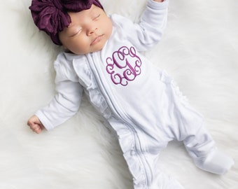 May include: A white baby sleeper with a ruffled trim and a purple monogram on the front. The sleeper is worn by a baby wearing a purple bow headband.