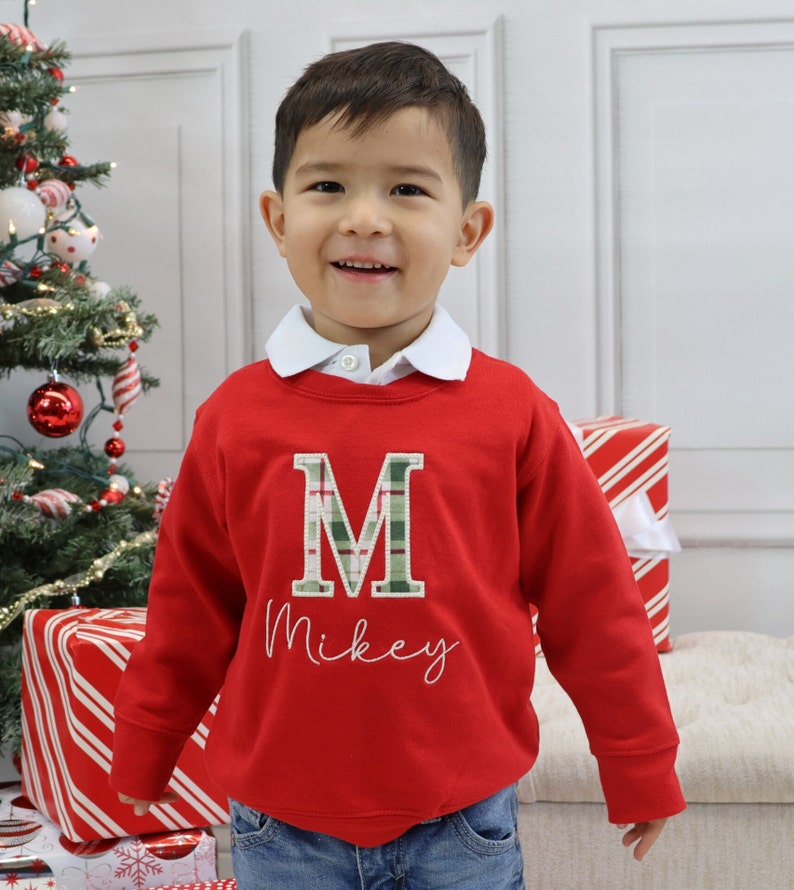 May include: A red sweatshirt with a white collar, featuring a green and white plaid letter "M" and the name "Mikey" in cursive. The sweatshirt is worn by a child, with a Christmas tree and wrapped gifts in the background.