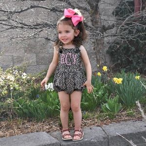 May include: A young girl wearing a leopard print romper with the word "BOY" embroidered on the front. She is wearing a pink bow in her hair and brown sandals.