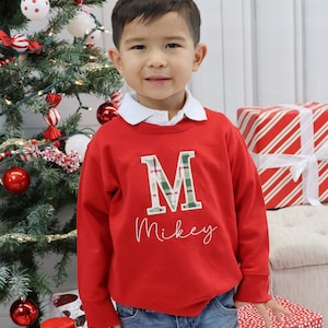 May include: A young child wearing a red sweatshirt with a white collar. The sweatshirt features a plaid "M" and the name "Mikey" embroidered in white. The child is standing in front of a Christmas tree and wrapped gifts.
