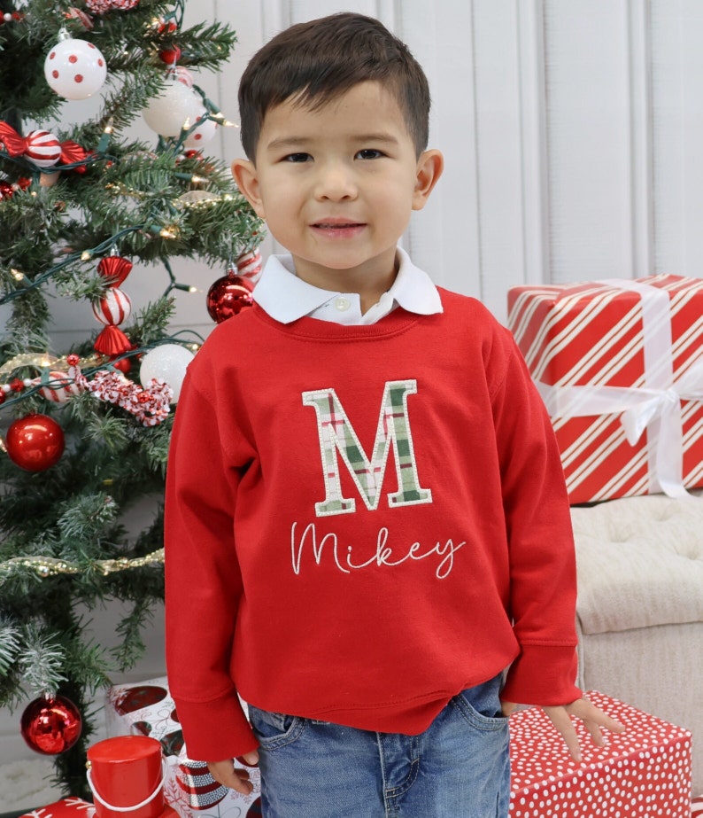 May include: A young child wearing a red sweatshirt with a white collar. The sweatshirt features a plaid "M" and the name "Mikey" embroidered in white. The child is standing in front of a Christmas tree and wrapped gifts.