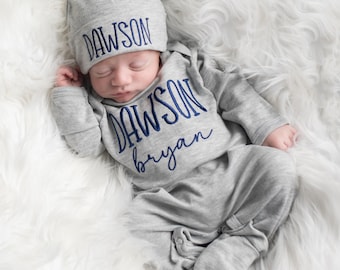May include: A newborn baby wearing a gray sleepsuit and matching cap, both embroidered with the name "DAWSON" in navy blue. The sleepsuit also has the name "bryan" embroidered in navy blue. The baby is lying on a fluffy white surface.