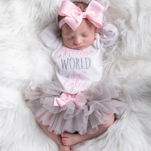 K&ouml;nnte beinhalten: Ein Baby M&auml;dchen tr&auml;gt einen wei&szlig;en Strampler mit dem Text "Hello World I'm Ezlee" in rosa und grauer Schrift. Sie tr&auml;gt ein rosa Schleifenhaarband und einen grauen T&uuml;llrock mit einer rosa Schleife.