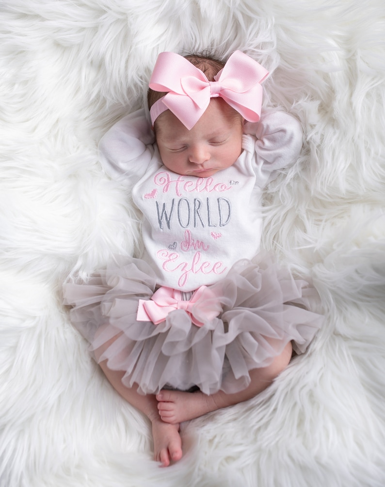 K&ouml;nnte beinhalten: Ein Baby M&auml;dchen tr&auml;gt einen wei&szlig;en Strampler mit dem Text "Hello World I'm Ezlee" in rosa und grauer Schrift. Sie tr&auml;gt ein rosa Schleifenhaarband und einen grauen T&uuml;llrock mit einer rosa Schleife.