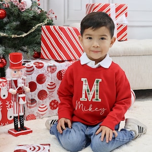 May include: A young child wearing a red sweatshirt with a green plaid "M" and the name "Mikey" embroidered on it. The child is sitting on a white rug in front of wrapped gifts and a Christmas tree. A decorative nutcracker stands nearby.