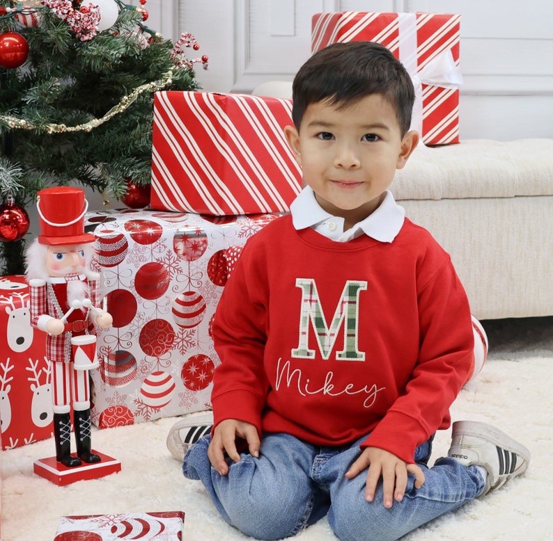 May include: A young child wearing a red sweatshirt with a green plaid "M" and the name "Mikey" embroidered on it. The child is sitting on a white rug in front of wrapped gifts and a Christmas tree. A decorative nutcracker stands nearby.