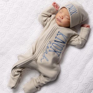 May include: A sleeping baby wearing a beige footed onesie and matching beanie. The onesie and hat are embroidered with the name "KNOX Noah" in blue script. The baby is lying on a white, textured blanket.