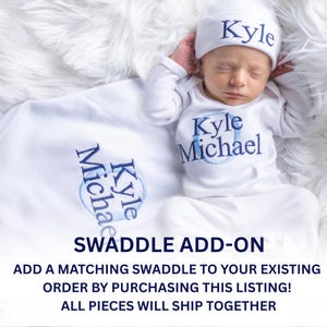 May include: A white long-sleeved baby bodysuit and matching beanie set featuring the embroidered name Kyle Michael in blue thread. A white swaddle blanket with the same blue embroidered name is included. Text reads: SWADDLE ADD-ON.