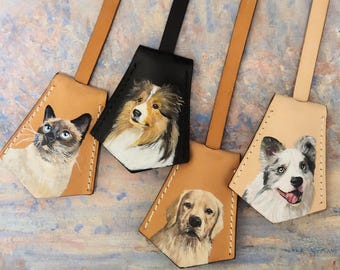 May include: Four leather keychains with painted pet portraits. The keychains feature a Siamese cat, a Collie, a Golden Retriever, and a Border Collie. Each keychain has a leather strap and a stitched border. The background is a colorful, abstract painting.