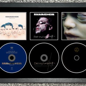 May include: A black framed display featuring three Rammstein album covers and three corresponding CDs. The top row shows Herzeleid, Sehnsucht, and Mutter artwork. The bottom row contains a blue CD, a black CD with text, and a black CD with a logo.