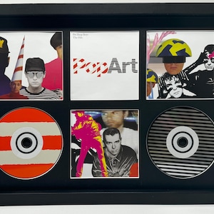 Puede incluir: Obra de arte enmarcada con portadas de álbumes y CD. La fila superior muestra imágenes de miembros de la banda y el texto "PopArt". La fila inferior muestra CD con diseños a rayas en naranja, blanco y negro. El marco es negro.