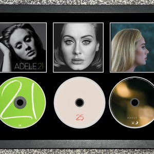 May include: A black framed display featuring three square photographic prints of Adele and three circular compact discs. The left disc is bright green with the number 21, the center disc is off-white with the number 25, and the right disc is dark.