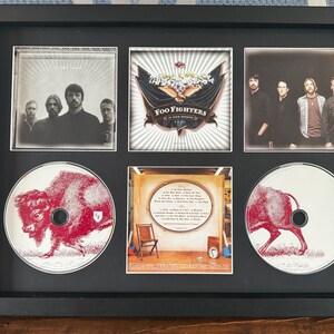 May include: Framed Foo Fighters memorabilia featuring album art and band photos. The frame includes images of the band members, album covers, and CDs with a red bison illustration. The artwork is displayed in a black frame.