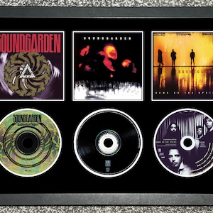 May include: Framed Soundgarden album art and CDs. The top row features album covers with the band's name in red. The bottom row displays three CDs. The frame is black, and the background is also black.