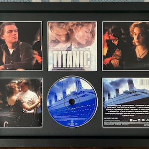 May include: A black framed display of Titanic movie memorabilia. The frame contains five film stills, a CD, and a CD insert. The CD is blue with a picture of the Titanic. The images feature actors Leonardo DiCaprio and Kate Winslet.
