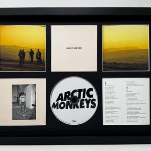 May include: Framed Arctic Monkeys album art display. The black frame holds a CD, album cover art, and lyric sheets. The CD is white with the band's name. The album cover art features a sunset landscape and band members.