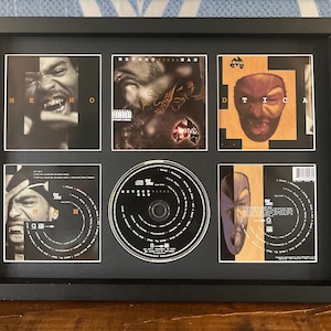 Puede incluir: Exhibición enmarcada en negro con un CD y portadas de álbumes de Method Man. El marco contiene un CD, cuatro portadas de álbumes y una imagen central. Las portadas presentan retratos y diseños gráficos, con el nombre del artista visible.