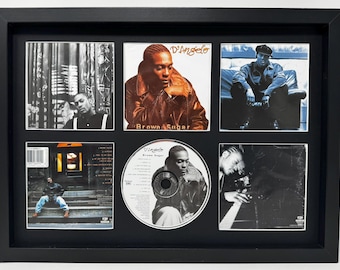 May include: Framed artwork featuring images of D'Angelo. The black frame holds six images, including CD art and album covers. The central CD has the album title "Brown Sugar". The images are in black and white and color.