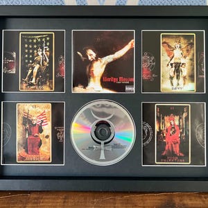 Puede incluir: Enmarcado con recuerdos de Marilyn Manson, con un CD y grabados artísticos estilo tarot. El CD está centrado, rodeado de imágenes con temas góticos y religiosos. El marco es negro.