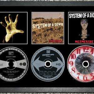 May include: A framed display featuring System of a Down album covers and CDs. The album covers are for "Toxicity" and "Mezmerize." The CDs have album art and text. The frame is black and the background is grey.
