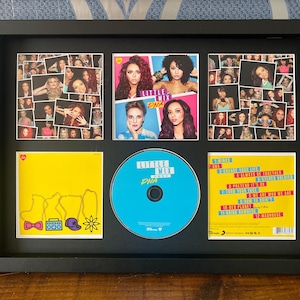May include: A framed Little Mix "DNA" album display. The black frame contains a CD, album art, and photo collages. The CD is blue, and the album art is yellow and blue. The photo collages feature multiple images of the band members.
