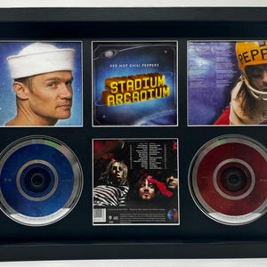May include: Framed music memorabilia featuring images of musicians and album art. Includes a portrait of a man in a sailor hat, album covers, and two CDs. The frame is black.