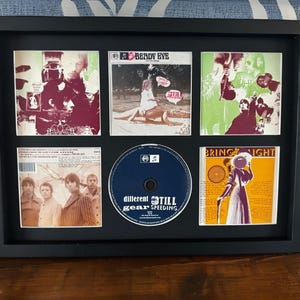 May include: A framed collection of music album covers and a CD. The frame is black and holds six album covers with various designs and colors. The CD is blue with the text "different STILL gear SPEEDING".