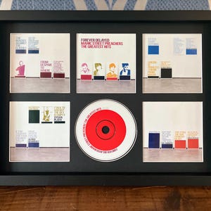 Puede incluir: Una exhibición enmarcada en negro con portadas de álbumes y un CD de la banda Manic Street Preachers. La exhibición incluye portadas de álbumes con texto y un CD rojo en el centro. El texto incluye "Forever Delayed".