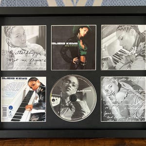 May include: A framed collection featuring images of Alicia Keys. The frame holds multiple small photos and a CD. The images are primarily black and white, with one color photo. The frame is black and rectangular.