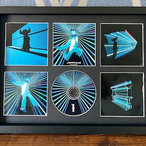 May include: Framed Jamiroquai album art and CD. The artwork features a silhouette figure with a horned headdress and geometric designs in shades of blue and black. The CD is centered, surrounded by six smaller images in a black frame.