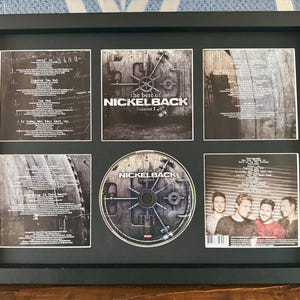 Może przedstawiać: Oprawiona prezentacja albumu "The Best Of Volume 1" zespołu Nickelback. Czarna ramka zawiera płytę CD i sześć paneli z okładkami albumu. Okładka albumu zawiera nazwę zespołu, tytuł albumu i tytuły piosenek.