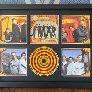 Op de afbeelding: Zwart ingelijste collage met afbeeldingen van de boyband *NSYNC. De collage bevat zes kleine foto's en een cd met een bordeauxrood en geel schietschijf-ontwerp.