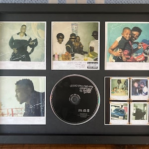 May include: A black framed artwork with a CD and several Polaroid-style prints. The prints show various images of people and scenes, with the CD in the centre. The CD has the text "good kid, m.A.A.d city" by Kendrick Lamar.