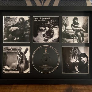 May include: Black framed Jamiroquai "Dynamite" album art and CD. The frame contains five black and white photos of the band and album art, with a CD in the center. The frame is rectangular and sits on a wooden surface.