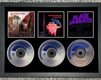 May include: Framed Black Sabbath album art and CDs. The top row features album covers for "Black Sabbath," "Paranoid," and "Master of Reality." Below are three CDs with blue and silver designs. The frame is black.