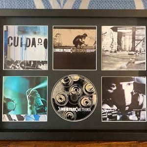 Può includere: Cornice nera contenente opere d'arte dell'album "Meteora" dei Linkin Park e CD. La cornice contiene sei immagini, tra cui la copertina dell'album e il CD. Le immagini presentano graffiti, vernice spray e ritratti dei membri della band. Il CD riporta il nome della band e il titolo dell'album.