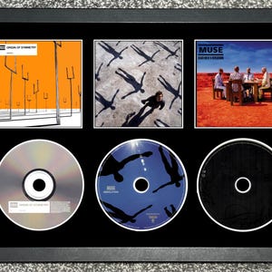 Puede incluir: Obra de arte enmarcada con tres portadas de álbumes y tres CD de la banda Muse. Las portadas de los álbumes tienen fondos naranja, gris y rojo. Los CD son plateados, azules y negros. El marco es negro.