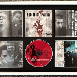 May include: A framed display of the Linkin Park Hybrid Theory album, featuring a white frame and black matting. It includes the album cover art, a red compact disc with track listings, and various band member portraits and group photographs.