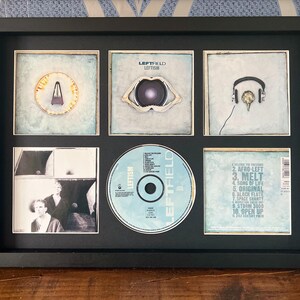 May include: A black framed display of Leftfield's album art and CD. The frame contains six images, including the CD and album covers with the text "LEFTISM". The album art features abstract designs and text. The CD is light blue.