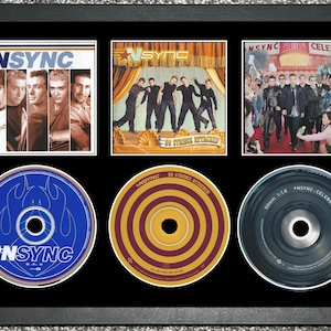 May include: Framed display featuring three album covers and three CDs by *NSYNC. The album covers showcase the band members, and the CDs have colorful designs. The frame is black, and the background is a light gray.