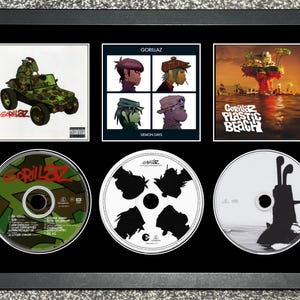 Puede incluir: Obra de arte enmarcada que presenta tres portadas de álbumes y tres CD de la banda Gorillaz. Las portadas de los álbumes incluyen "Gorillaz", "Demon Days" y "Plastic Beach". Los CD tienen varios diseños, incluyendo camuflaje y siluetas.