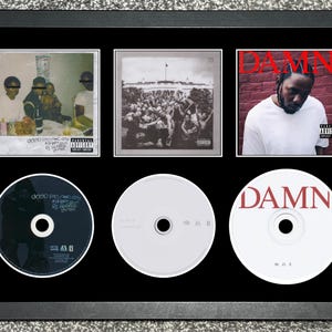 Puede incluir: Obra de arte enmarcada con tres portadas de álbumes y tres CD. Las portadas de los álbumes muestran imágenes de personas, una multitud y un retrato. Los CD son blancos y negros. La palabra "DAMN" es visible en una portada y un CD.